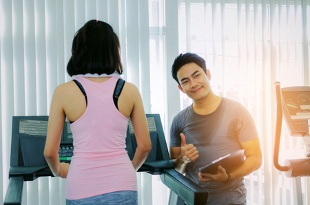muscular handsome personal trainer man with clipboard showing thumbs up and young asian woman slim body exercising on treadmill machine in fitness gym, work out, bodybuilder, healthy and sport conceptの写真素材