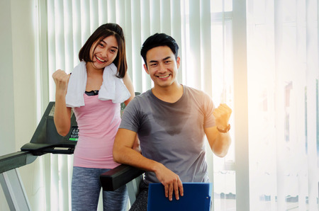 young happy smile asian woman slim body with towel and muscular personal trainer man with clipboard showing cheer up hand while resting after workout in fitness gym, healthy, exercise, sport conceptの写真素材