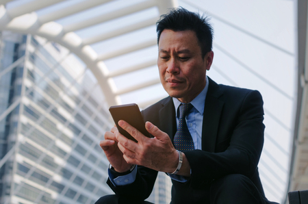 medium aged senior business man wearing modern black suit using mobile smart phone and briefcase sitting on walkway in city, internet network connection, technology communication and financial conceptの写真素材