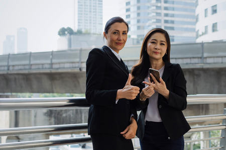 two business asian and caucasian woman with mobile phone and giving thumbs up as like sign together in the city, successful, support, meeting, partner, teamwork, community and connection conceptの写真素材