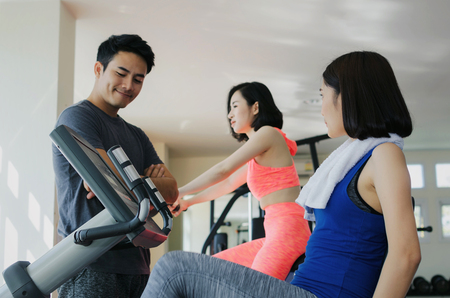 asian young woman slim body exercising on bicycle machine with young man personal trainer in fitness gym, bodybuilder, healthy lifestyle, fitness, workout and sport training concept, soft focusの写真素材