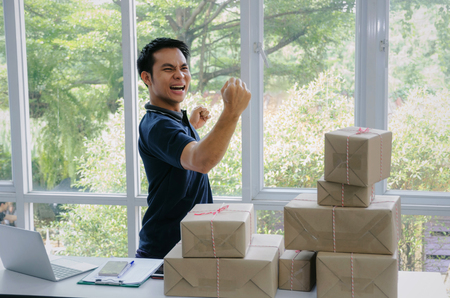 Start up. young man happy after new order from customer with laptop computer, mobile phone, delivery packaging box on table, small business owner working at home office, SME, shopping online conceptの写真素材
