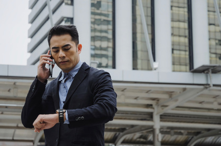 young smart asian business man wearing modern black suit looking at his watch and making phone call with mobile smart phone in city background, network connection, technology communication conceptの写真素材