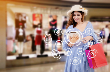 asian woman watching special offer in tablet and holding shopping bag while standing at new collection clothes shop in department store shopping mall with icon product diagram, shopping online conceptの写真素材
