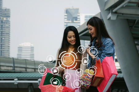 two beautiful asian young woman watching special offer in mobile phone and holding shopping bag in big city with icon product diagram, technology, summer sale, payment and shopping online conceptの写真素材