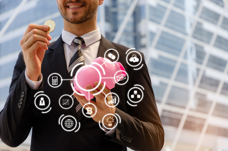 caucasian smart business man in black suit holding piggy bank and golden Bitcoin on modern city with graphic icon diagram, digital technology, virtual currency, financial, investment conceptの写真素材