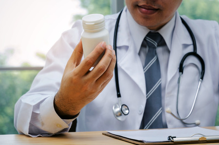 doctor or pharmacist with stethoscope on neck sitting at worktable with clipboard, laptop and holding drug bottle in hospital or clinic, medical technology, disease treatment, health care conceptの写真素材
