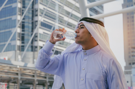 young handsome Arabian man in white suit standing and drinking natural water from bottle for refresh yourself with modern city background, lifestyle, healthy, summer conceptの写真素材