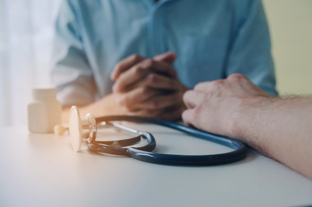 close up of hand of doctor and suffering patient sitting with stethoscope and bottle of pills on desk in hospital, encouragement, health care, medical, medicine, pharmacy and insurance conceptの写真素材