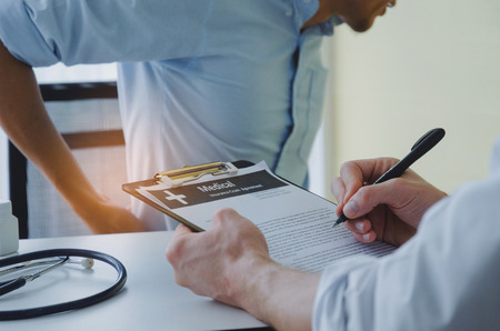 young business man suffering from backache meet doctor writing prescription on clipboard with stethoscope on desk in hospital, office syndrome, health care, medical, medicine, pharmacy conceptの写真素材
