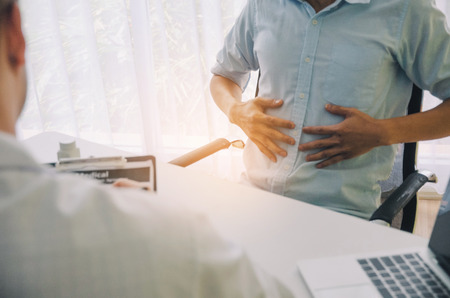 young man suffering from stomachache meet doctor writing prescription on clipboard with laptop on desk in hospital, office syndrome, health care, medical, medicine, pharmacy conceptの写真素材