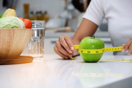 close up young woman hand holding measuring tape around fresh green apple on white table in kitchen, healthy food, clean eating, dieting, health care, weigh loss and vegetarian food conceptの写真素材