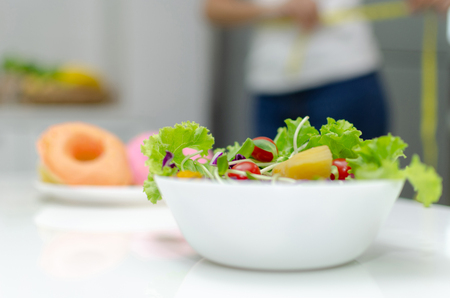 healthy fresh mix vegetable salad in white bowl with delicious glazed donuts in background on desk in kitchen interior at house, vegetarian, lifestyle, vegan, good healthy, diet food and drink conceptの写真素材