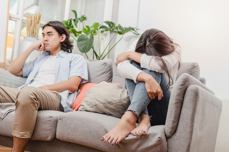 sadness young asian wife having quarrel and sitting on sofa after fight with husband behind her in house interior together, upset couple, love, divorce couple, family issues and relationship conceptの写真素材