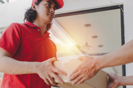 home delivery service handsome man in red uniform handing parcel boxes to recipient and young man customer accepting parcel post box from courier at home, express delivery and online shopping conceptの写真素材