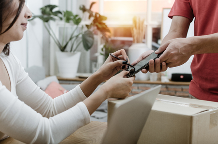 young woman customer appending signature in digital mobile phone receiving parcel post box from courier with home delivery service man in red uniform at home, express delivery, online shopping conceptの写真素材