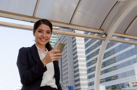 beautiful smiling young asian business woman wearing modern black suit playing social network with mobile phone in building city background, network technology, internet and digital conceptの写真素材