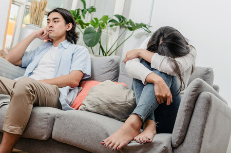 sadness young asian wife having quarrel and sitting on sofa after fight with husband behind her in house interior together, upset couple, love, divorce couple, family issues and relationship conceptの写真素材
