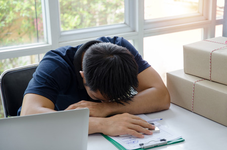 Start up. young man sleeping and tired during working on desk with laptop computer, clipboard and delivery parcel packaging box on table, small business owner at home office, shipping and SME conceptの写真素材