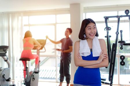 young happy smiling asian woman slim body with towel crossing arms in fitness gym with people on machine bicycle background, bodybuilder, healthy lifestyle, exercise, workout, sport training conceptの写真素材