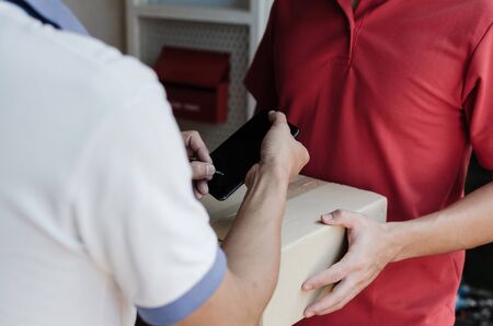 young man customer appending signature in digital mobile phone receiving parcel post box from courier with home delivery service man smiling face in red uniform at home, express delivery conceptの写真素材