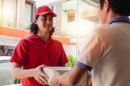 young man customer receiving parcel post box from courier with delivery service man smiling face in red uniform at home, cargo shipping, express delivery service, online shopping and logistics conceptの写真素材