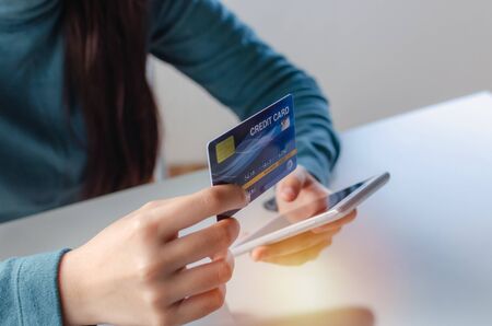 young woman entering security code with mobile smart phone and paying credit card on desk at home...