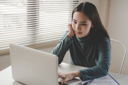 pretty young asian woman typing working and using laptop and document on desk at home office,...