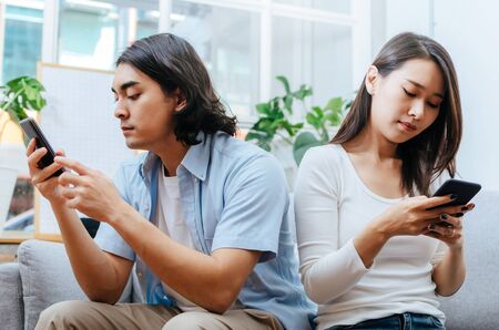 asian young couple man and woman absorbed and focused into mobile phone and sitting on sofa in...