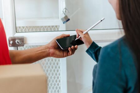 woman customer using pen signature on digital mobile phone with delivery service man in red uniform for receiving parcel post box from courier at home, express delivery and shopping online conceptの写真素材
