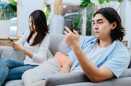 asian young couple man and woman absorbed and focused into mobile phone and sitting on sofa in living room at home, social media addiction, upset couple, family issues and relationship conceptの写真素材