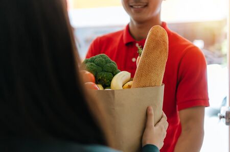 smart food delivery service man in red uniform smiling and sending fresh food set to young woman customer receiving order from courier at home, express delivery, food delivery, online shopping conceptの写真素材