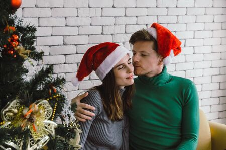 young caucasian sweet couple with red santa hat enjoy and kissing his girlfriend on sofa with...