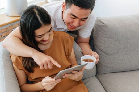 young asian love couple drinking coffee and using mobile tablet, watching movie online on sofa at home office, internet technology, social media, online booking, entertainment, shopping online conceptの写真素材