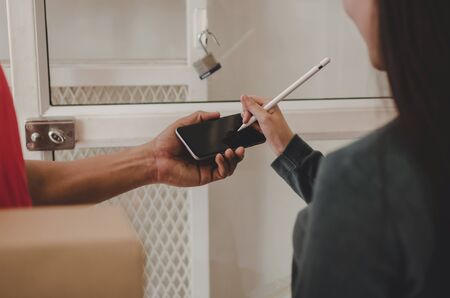 home delivery service man in red uniform and young woman customer appending signature in digital mobile phone receiving parcel post box from courier at home, express delivery, online shopping conceptの写真素材