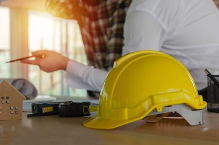 yellow safety helmet on workplace desk with construction worker team meeting and planning about building plan in office center at construction site, real estate, engineering and contractor conceptの写真素材