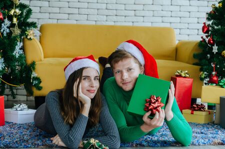 Merry christmas. young caucasian love couple with red santa hat showing gift box and laying together on fall with christmas tree background at home, holiday family, happy new year, festival conceptの写真素材