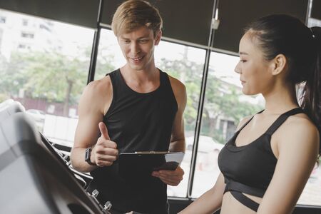 muscular handsome caucasian personal trainer man showing thumbs up with young asian woman slim body exercising his legs cardio running on treadmill in fitness gym for good healthy, sport conceptの写真素材