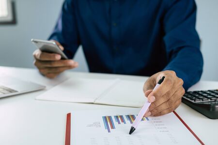 business man in blue shirt using mobile phone calculating about finance with document report on desk at home, self quarantine, work from home, marketing, business financial, digital technology conceptの写真素材