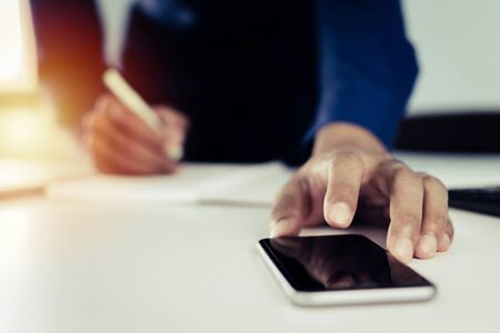 young man in blue shirt hand working on digital mobile phone and writing about finance with document report on desk at home office, work from home, digital marketing and financial business conceptの写真素材