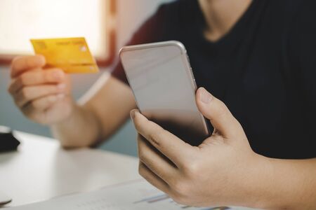 young man entering holding mobile phone and paying with mock up credit card on desk at home office, digital marketing, shopping online, electronic pay, online payment and digital technology conceptの写真素材