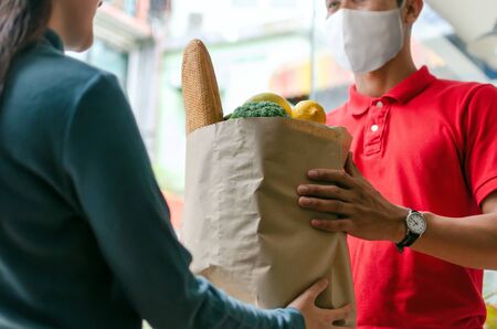 woman customer receiving fresh food set bag from food delivery service man with protection face mask in red uniform at door home, express delivery, quarantine, virus outbreak, food delivery conceptの写真素材