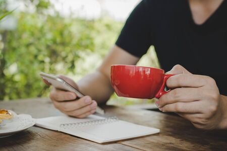 business man relaxed holding red coffee cup and working with digital mobile phone on desk at cafe, self quarantine, work from home, digital marketing, business finance, network technology conceptの写真素材