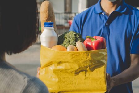 food delivery service man with protection face mask in blue uniform holding fresh food set bag to customer at door home, express delivery, quarantine, virus outbreak, takeaway food delivery conceptの写真素材