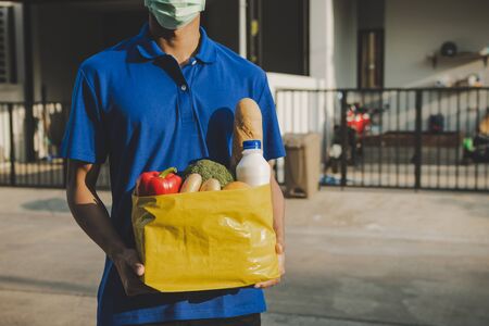handsome delivery man with protection face mask in blue uniform holding fresh food set bag waiting for customer at door home, healthy food, express delivery, food delivery, online shopping conceptの写真素材