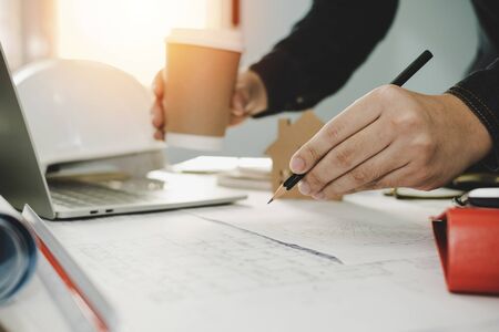 architect, engineer or interior designer hands drawing with pencil on blueprint and holding coffee cup on workplace desk in office center at construction site, construction and architectural conceptの写真素材