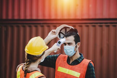 industrial worker woman temperature checking fever by digital thermometer with engineer man with cargo container in background at container cargo harbor, industrial, logistic import and export conceptの写真素材