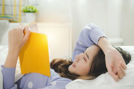 Stay Home. happy young asian pretty woman in casual clothing lying on bed and enjoying reading a book at morning in bedroom at home, joyful weekend, student, leisure, quarantine and lifestyle conceptの写真素材