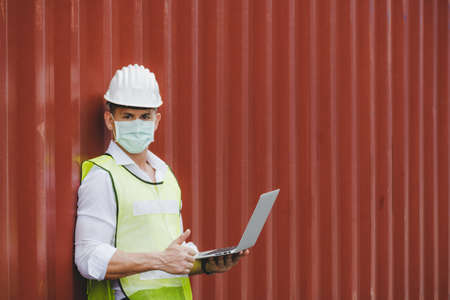 caucasian engineer control worker wearing protection face mask working on digital laptop computer with cargo container in background at cargo harbor, industrial, logistic import and export conceptの写真素材