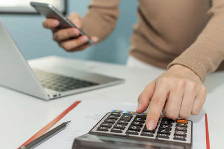 businesswoman hand calculate about financial cost bill with calculator and working on digital mobile phone and laptop computer on desk at home office, digital marketing, business finance conceptの写真素材